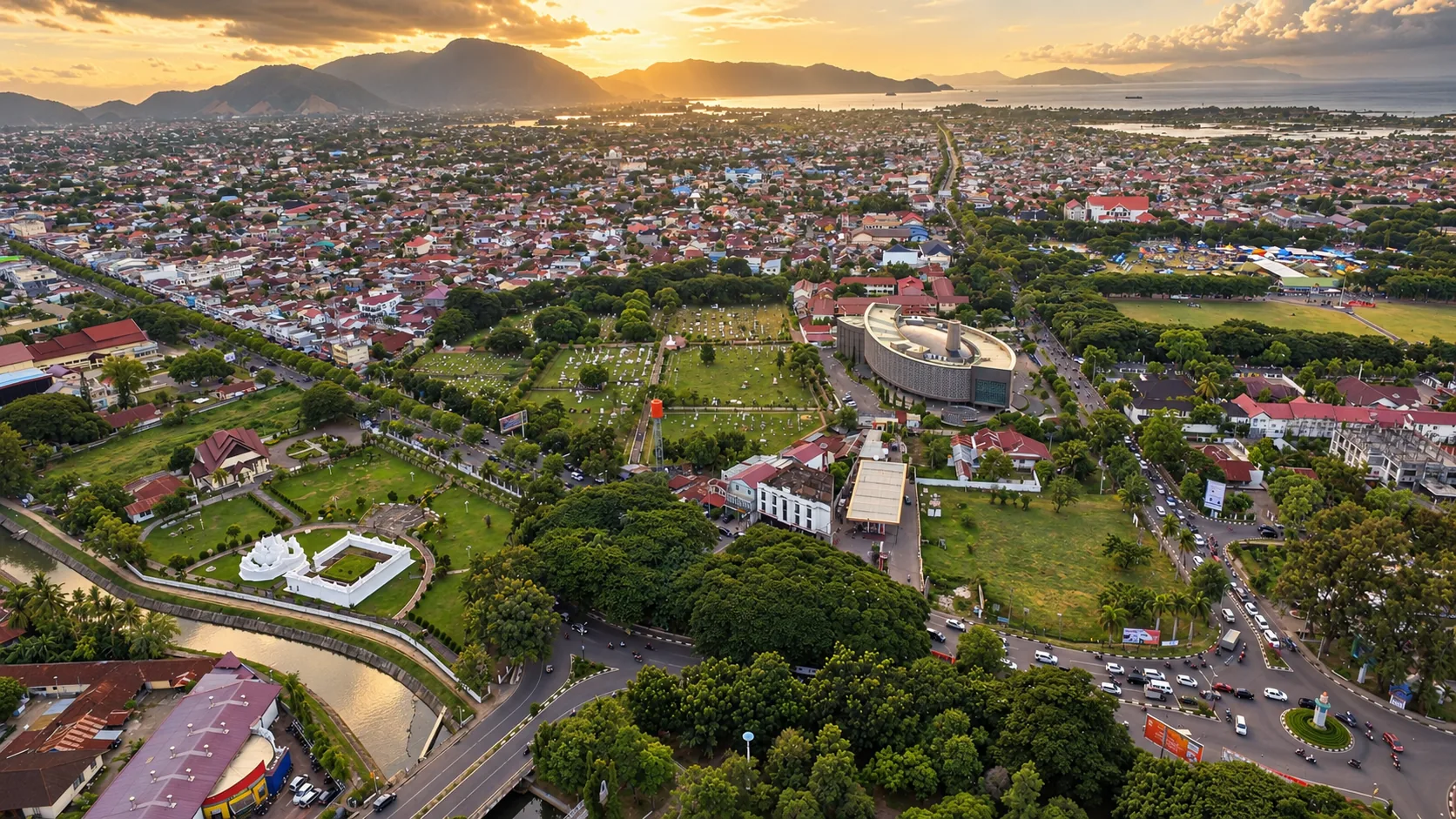 Aerial view of Banda Aceh at dusk: rooftops, green parks and the curved Aceh Tsunami Museum, with hills and the sea behind